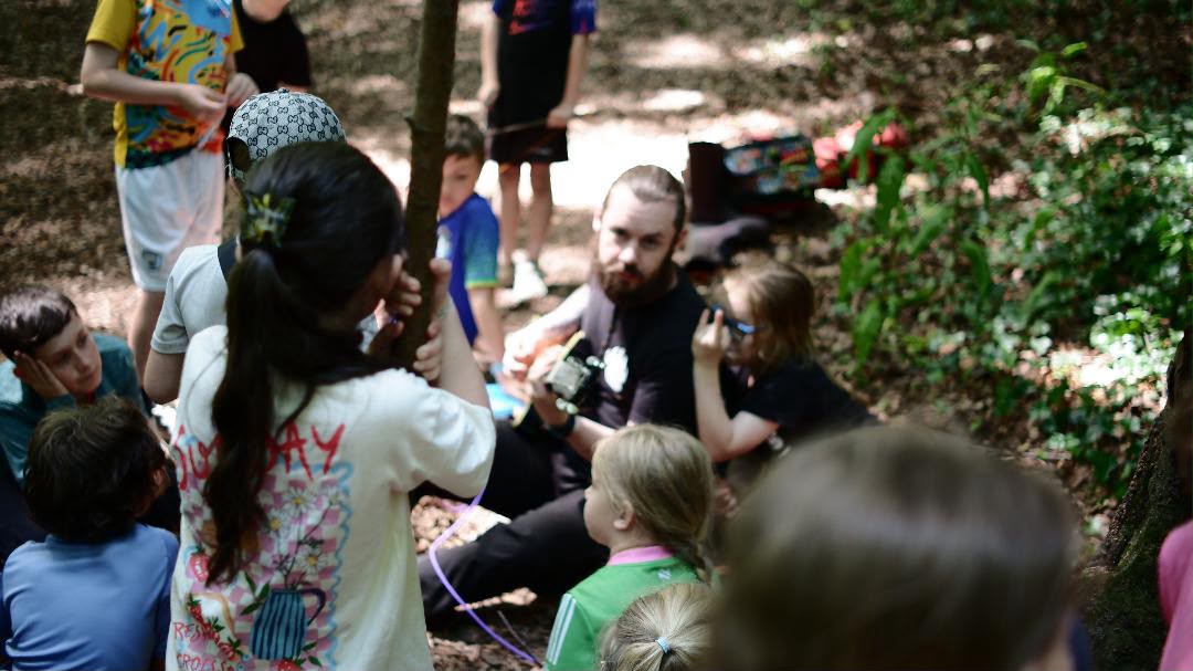 Camp leader Jack sitting on the woodland floor playing guitar, surrounded by children