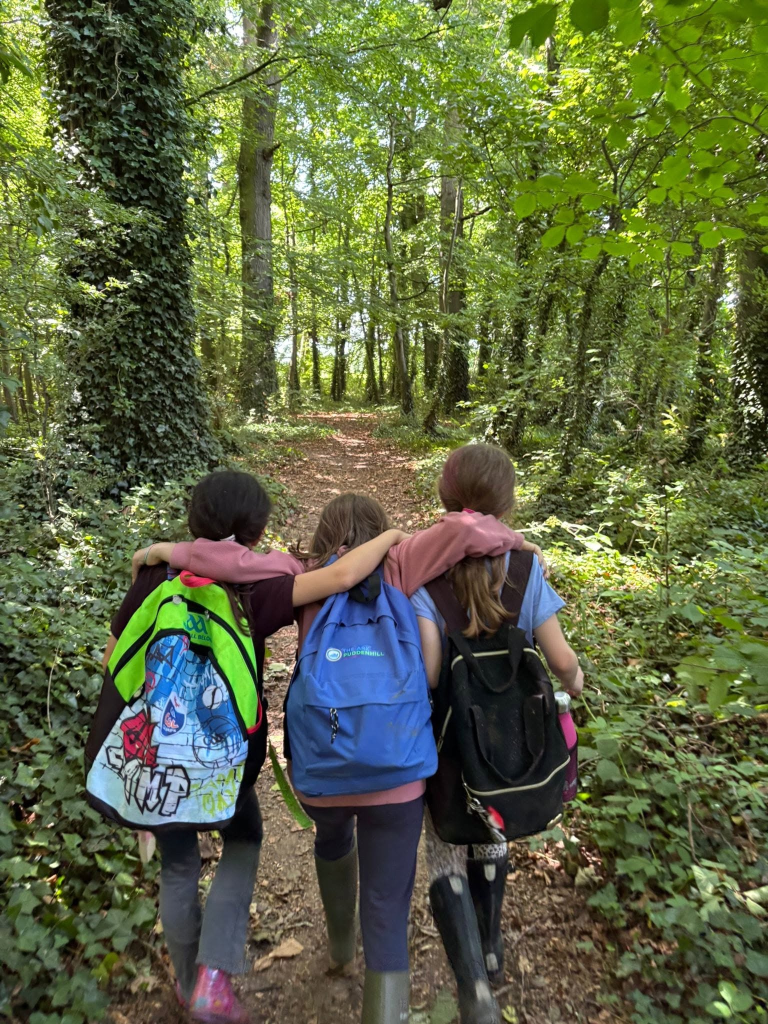 Three girls with backpacks walking arm in arm down a leafy woodland path, seen from behind