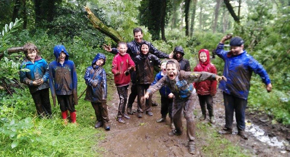 A group of children in muddy rain jackets laughing and pulling faces on a wet woodland trail with a camp leader